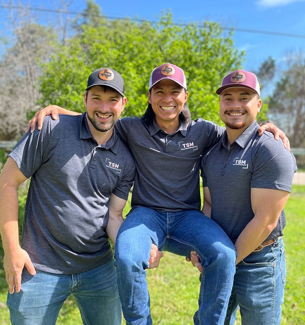 Three men in matching gray polo shirts and baseball caps featuring the TSM logo pose together outdoors.