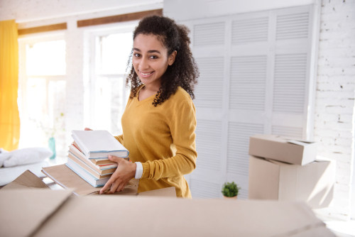 A smiling woman with curly hair, wearing a yellow shirt, holds a stack of books while unpacking boxes in a bright room.