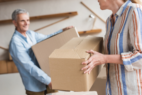 An elderly couple is shown carrying cardboard boxes. The man, with gray hair and a blue shirt, is smiling as he holds a box, while the woman, wearing a striped shirt, assists him.