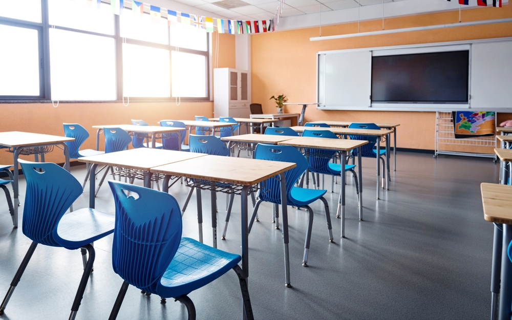 An empty classroom with desks and blue chairs, a whiteboard, and a large screen on the wall.