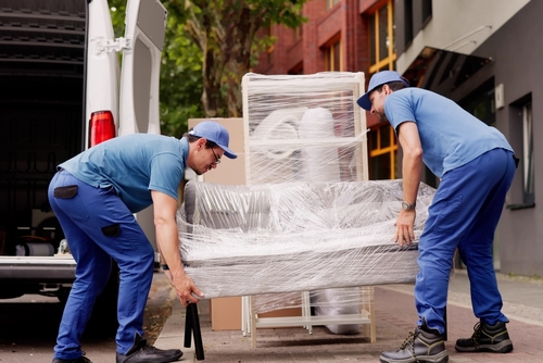 Two men in blue uniforms and caps unloading a wrapped couch from a van.