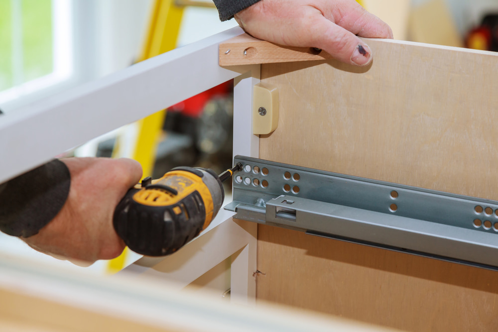 A person using a cordless drill to install a metal rail onto a wooden cabinet structure.